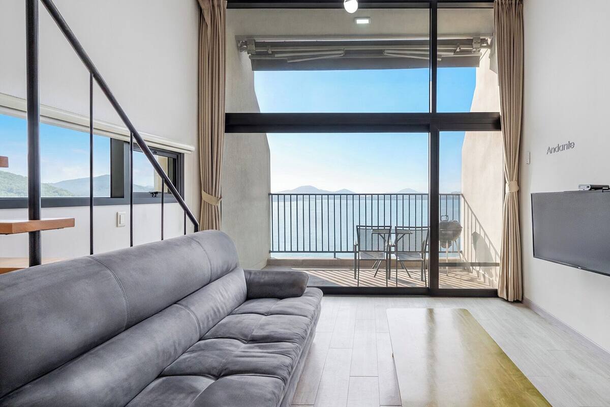 A modern living room with a grey sofa, a flat-screen TV, and a balcony with a view of the sea.