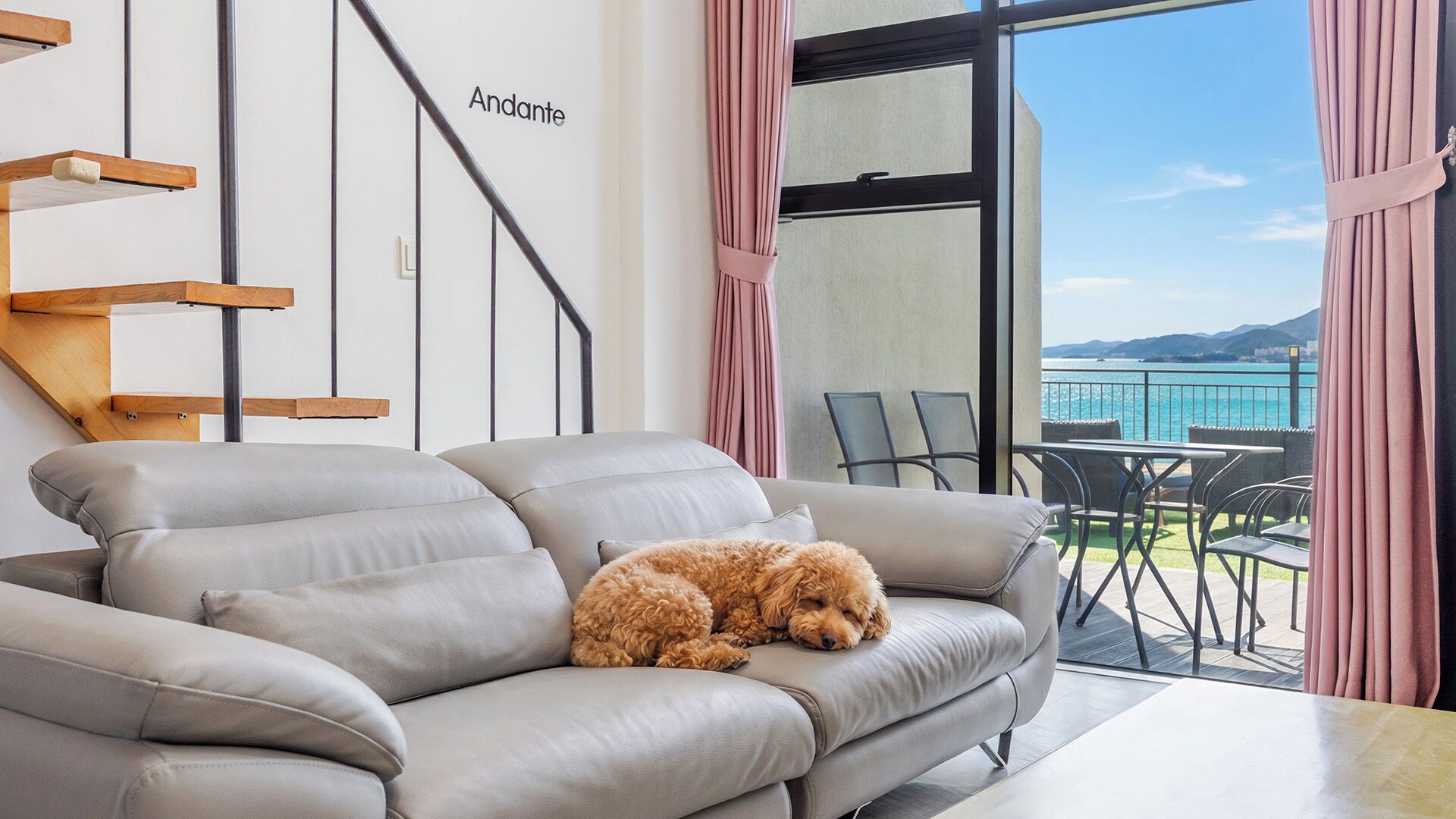 A modern living room with a sofa, a dog, a staircase, and a view of the sea.