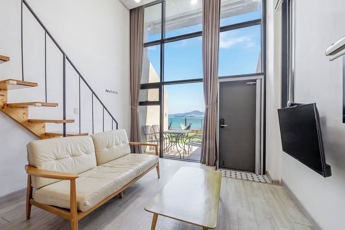 A modern interior with a sofa, a dining table, a TV, a staircase, and a large window with a view.