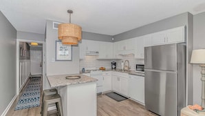A modern kitchen with white cabinets, a stainless steel refrigerator, a hanging wicker light fixture, and a small island with a countertop. - Marshside Inn 107 (Murrells Inlet)