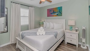 A bedroom with a bed, a ceiling fan, a painting, a nightstand, and a bedside table. - Marshside Inn 107 (Murrells Inlet)