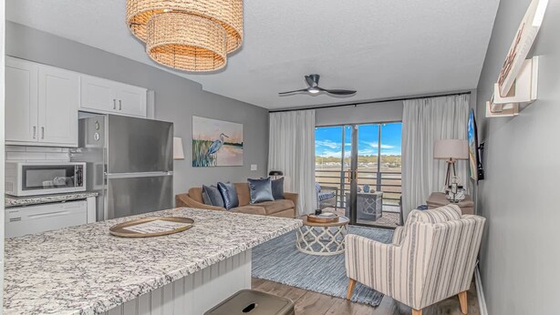 A modern kitchen with a large island, a dining area with striped chairs, and a view of the outdoors. - Marshside Inn 107 (Murrells Inlet)
