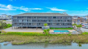 A large, modern building with a flat roof and multiple windows. In front, there is a swimming pool with lounge chairs and a small deck area. - Marshside Inn 107 (Murrells Inlet)
