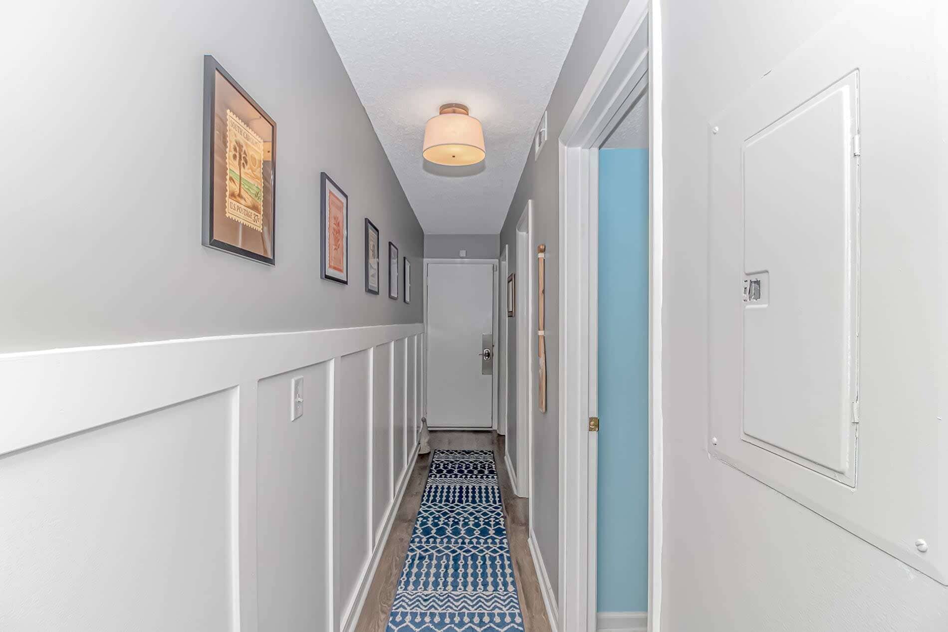 A narrow hallway with a patterned carpet, framed pictures on the walls, and a door with a light switch.