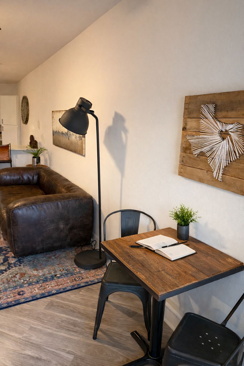 Une pièce comprenant un bureau en bois, une chaise noire, un canapé en cuir, un lampadaire, une œuvre d’art murale et une petite plante en pot.