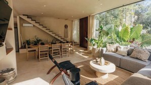 A modern living room with a staircase, a dining area, and a large window with plants. - Villa Selva | 4BR Villa w/ Private Pool & Rooftop (Tulum)
