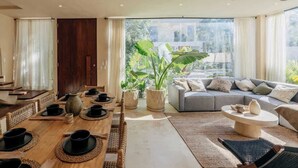 A modern living room with a dining area, large windows, and a view of greenery. - Villa Selva | 4BR Villa w/ Private Pool & Rooftop (Tulum)