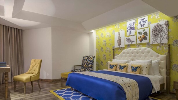 A bedroom with a bed, a chair, a desk, and a wall with floral artwork. - Mosaic Mussoorie (Dehradun)