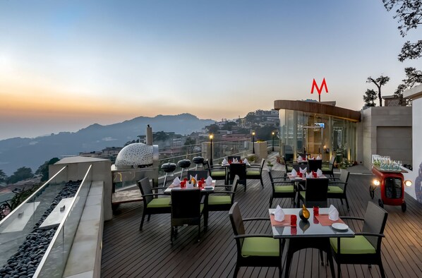 A rooftop restaurant with tables and chairs, a view of the city, and a mountain in the background. - Mosaic Mussoorie (Dehradun)