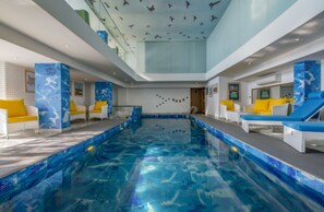 A large indoor swimming pool with a tiled floor, surrounded by white and yellow lounge chairs. - Mosaic Mussoorie (Dehradun)