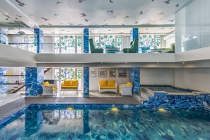 Ein mehrstöckiger, überdachter Hallenbad mit Wandfliesen aus Mosaik, einer Glasdecke mit Vogelkonturen und Sitzbereichen mit gelben Kissen.
