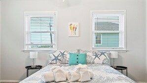 Een slaapkamer met een bed, twee nachtkastjes, een raam met zonwering, een wanddecoratie en een turquoise kussen.