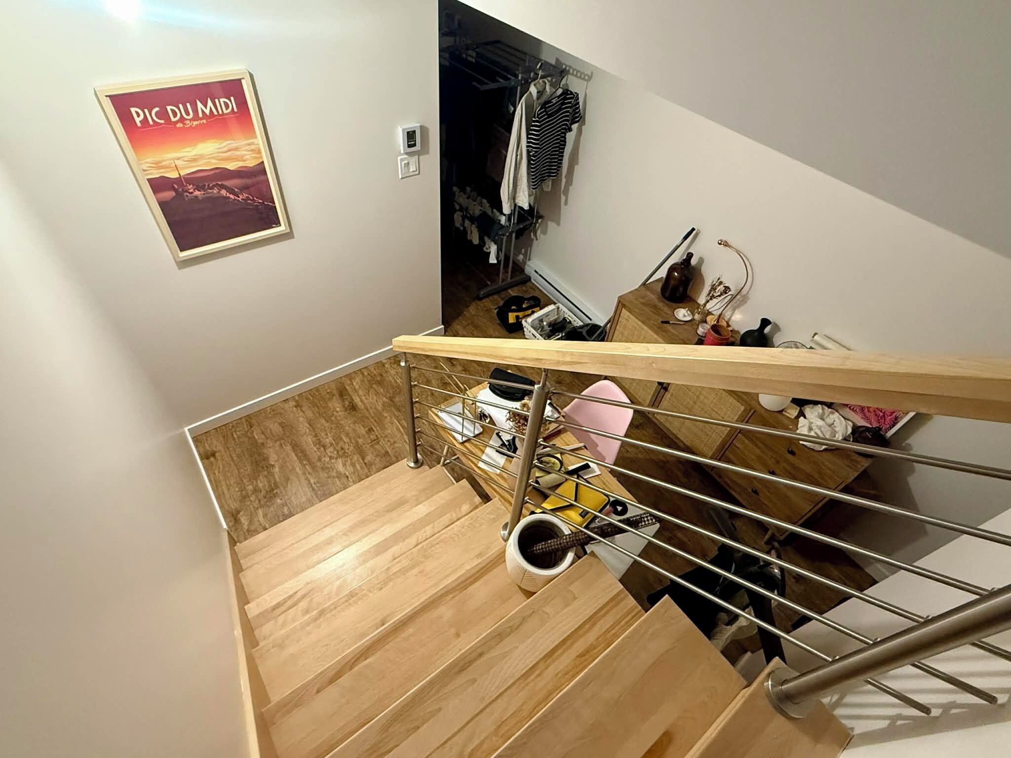 Un escalier avec des marches en bois et des rampes en métal mène à une pièce où se trouvent un bureau en bois, une chaise et une affiche accrochée au mur.