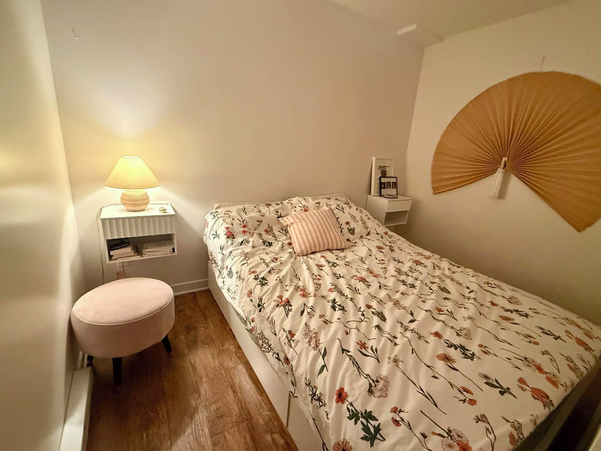 Un dormitorio con una cama, una mesita de noche con lámpara, un taburete pequeño y un ventilador fijado a la pared.