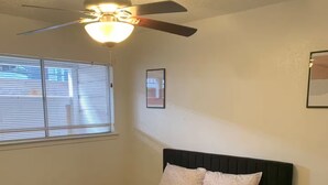 Un dormitorio con cama, ventilador de techo, ventana con persianas, mesita de noche y alfombra.