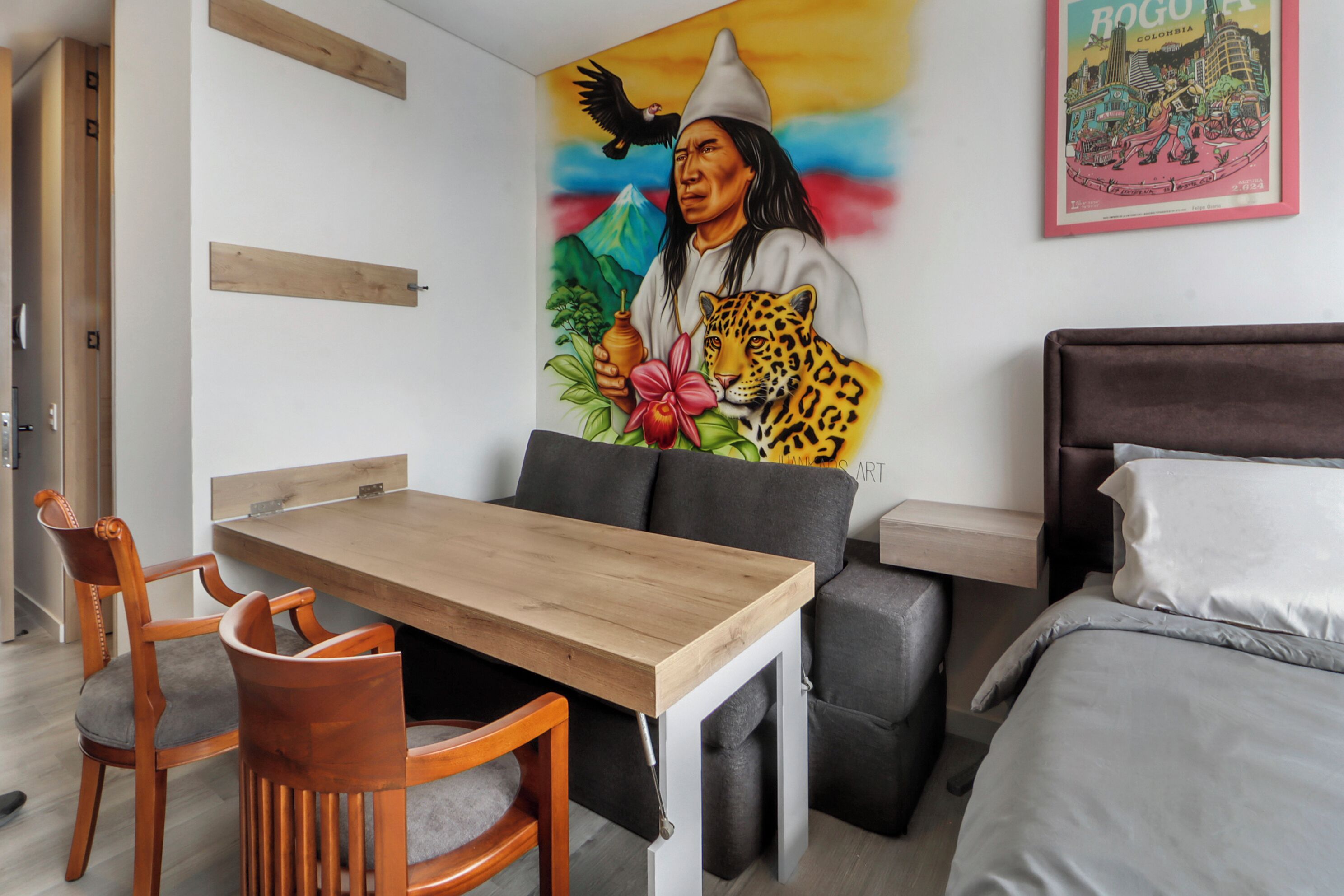 Une pièce comprenant une table à manger en bois, deux chaises, un lit avec une literie blanche, un canapé gris et une fresque colorée sur le mur.
