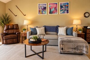 A living room with a brown leather recliner, a grey sofa with blue and white pillows, a round coffee table with a basket, and a clock on the wall. - 14th Fairway Retreat | Solar, Battery & EV Charger (Palm Desert)