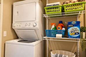 A laundry area with a front-loading washing machine, shelves with laundry detergent, and a basket. - 14th Fairway Retreat | Solar, Battery & EV Charger (Palm Desert)