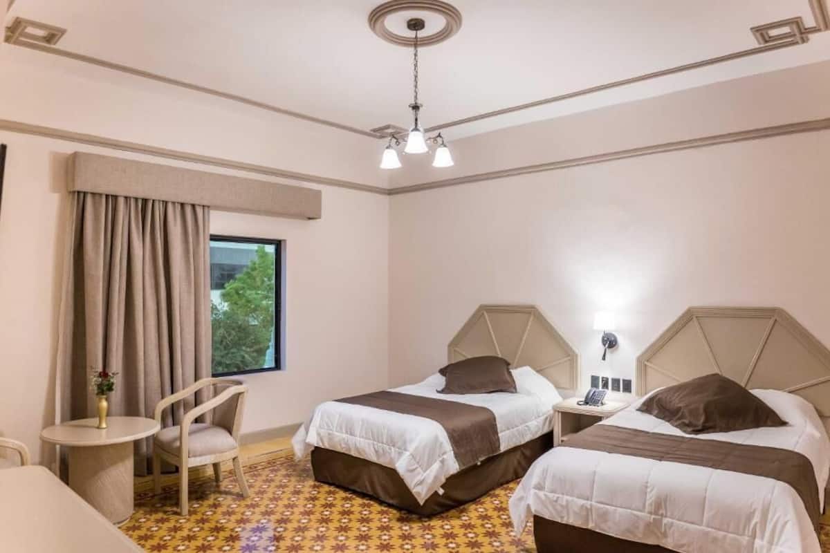 A hotel room with two beds, a small table, a chair, a window with curtains, and a ceiling light fixture.