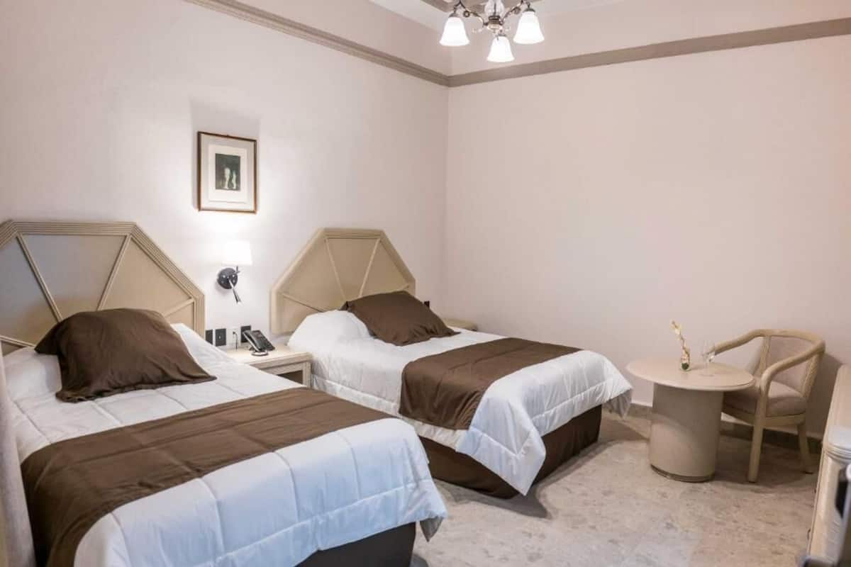 A hotel room with two beds, a bedside table with a lamp, a small round table with a chair, and a picture on the wall.