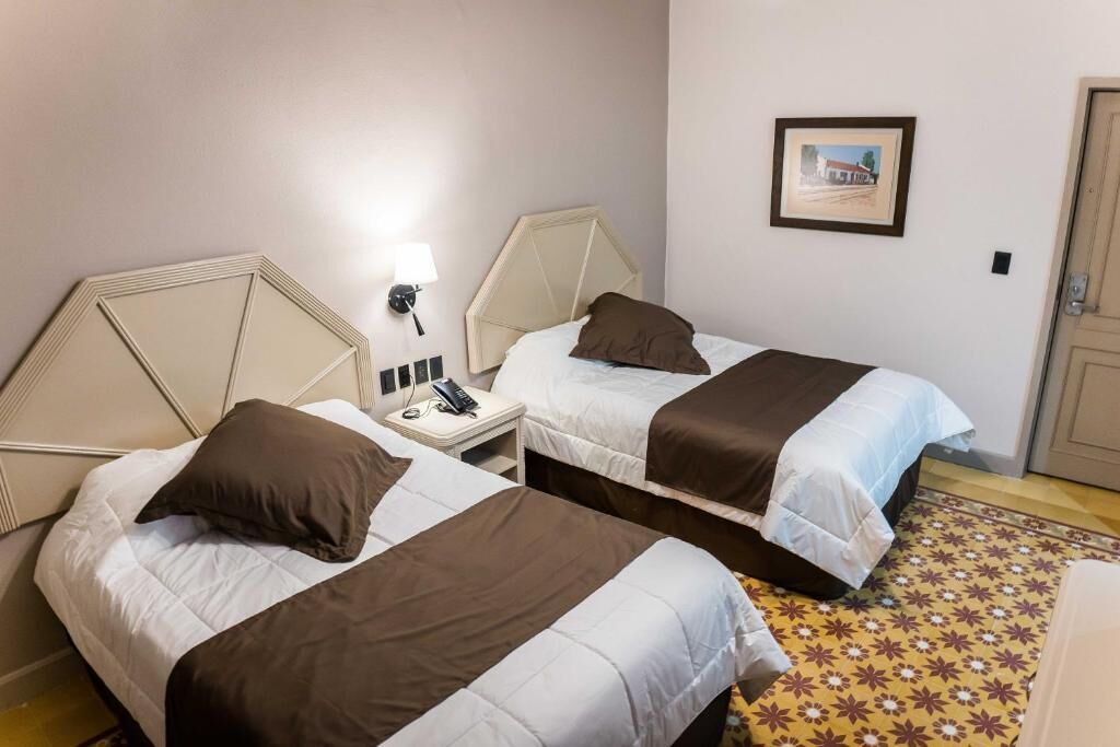A hotel room with two beds, a nightstand with a lamp, a phone, and a framed picture on the wall.