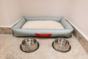 Una cama para mascotas con un asa roja y dos tazones metálicos sobre un suelo de baldosas.