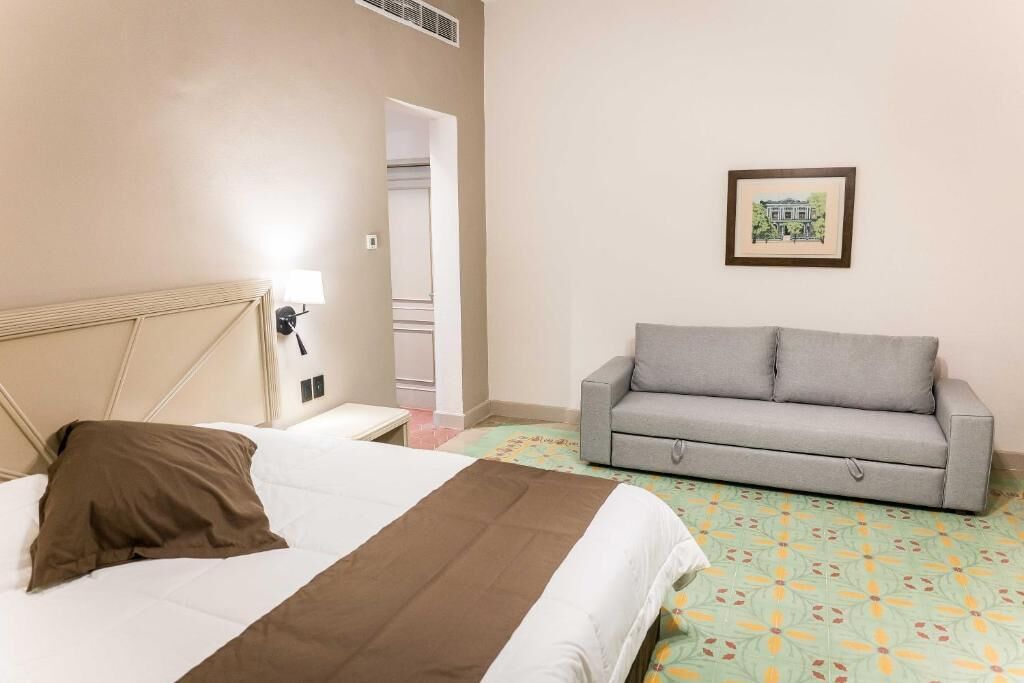 A hotel room with a bed, a grey sofa, a framed picture on the wall, and a door leading to a bathroom.