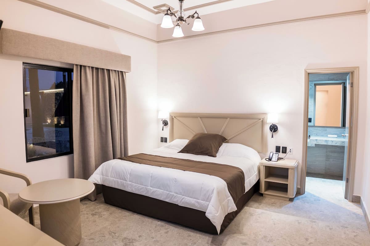 A hotel room with a bed, bedside table, lamp, and a view of the outside.