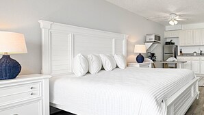 Une chambre avec un lit blanc, une table de chevet avec une lampe, une commode et un coin cuisine équipé de placards et d’appareils électroménagers.