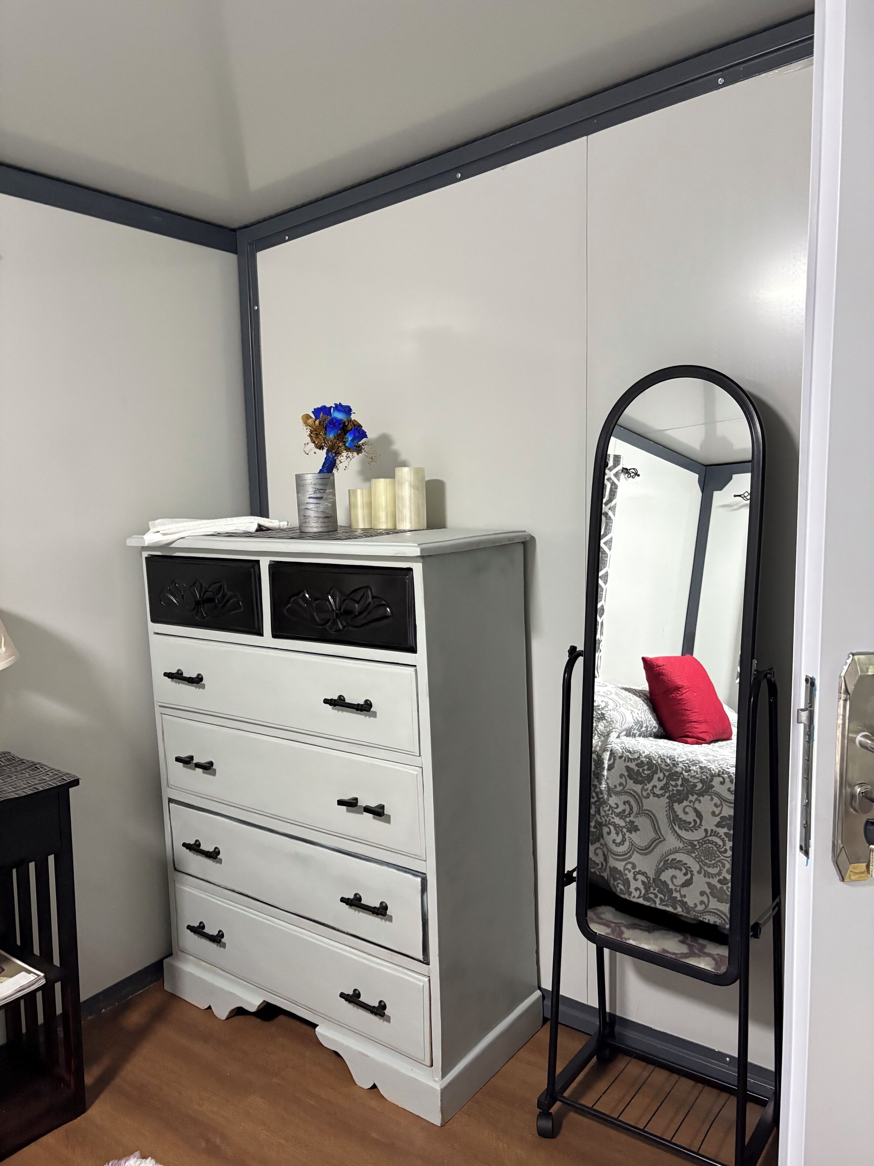A bedroom with a white dresser, a mirror, a bed with a patterned bedspread, and a small table with a lamp.