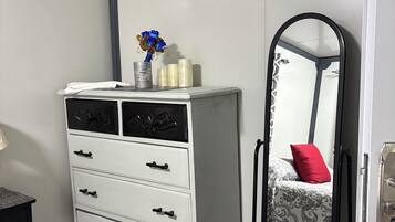 A bedroom with a white dresser, a mirror, a bed with a patterned bedspread, and a small table with a lamp.