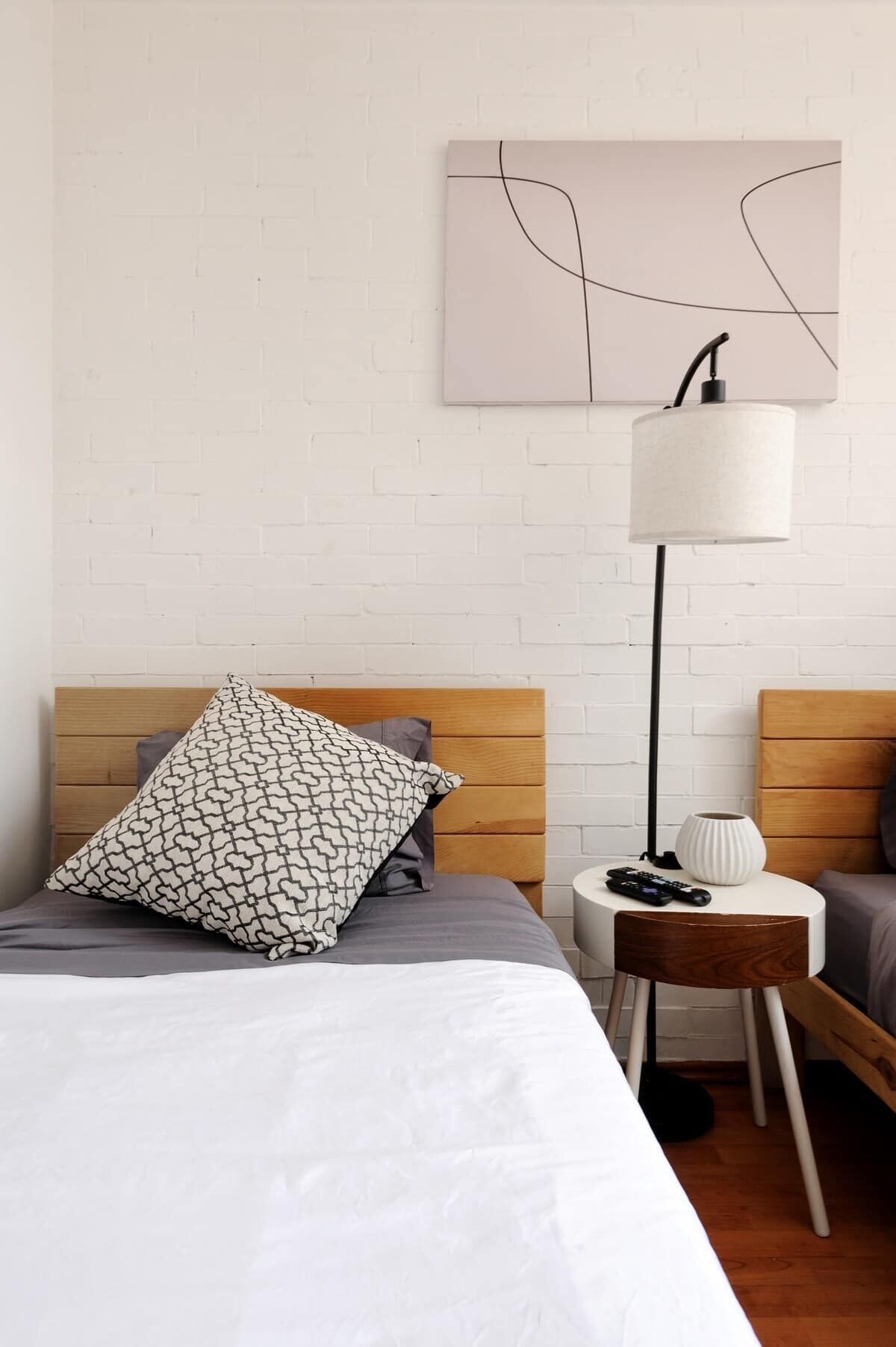 Une chambre d’hôtel avec un lit en bois, une lampe blanche, une petite table d’appoint et un mur en briques blanches.
