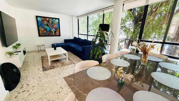 A modern living room with a large TV, blue sofa, dining table with white chairs, and a large window with a view of trees. - 4BR apartment | Private Balcony · Near Masaryk (Mexico City)