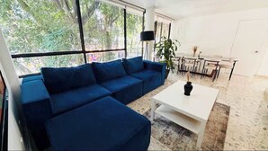 A modern living room with a sectional sofa, a dining table with chairs, a white coffee table, and a large window with a view of trees. - 4BR apartment | Private Balcony · Near Masaryk (Mexico City)