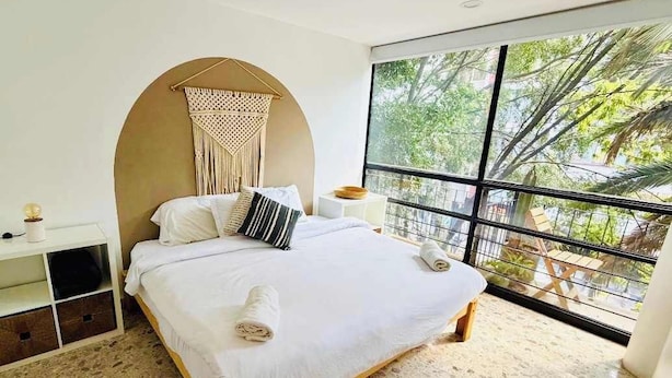 A bedroom with a bed, a bedside table, a wall-mounted macrame piece, and a large window with a view of trees. - Boutique Apt | Polanco - Masaryk (Mexico City)