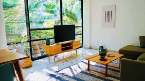 A living room with a TV on a wooden stand, a coffee table, a sofa, and a large window with a view of greenery. - Boutique Apt | Polanco - Masaryk (Mexico City)