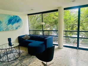 A modern living room with a blue sofa, a glass coffee table, and a black chair. There is a large window with a view of greenery outside. - PH in Polanco | Near Masaryk · 4BR · Balcony (Mexico City)