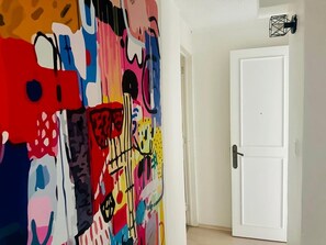 Huoneessa on värikäs, abstrakti seinämaalaus, valkoinen ovi ja musta seinään kiinnitetty valaisin.