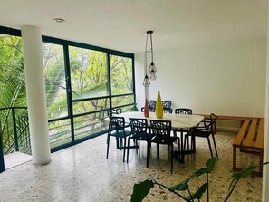 Ein Essbereich mit einem runden Tisch, Stühlen und einer Bank. Große Fenster bieten Blick auf Grünflächen.