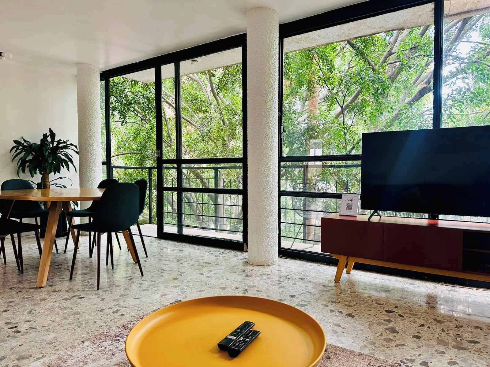 Une pièce avec une grande fenêtre, un téléviseur, une table à manger avec des chaises et un plateau jaune.