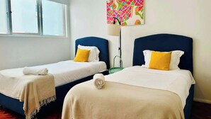 Habitación de hotel con dos camas, cada una con cabecero azul y ropa de cama blanca. Hay un cuadro abstracto y colorido en la pared sobre las camas.