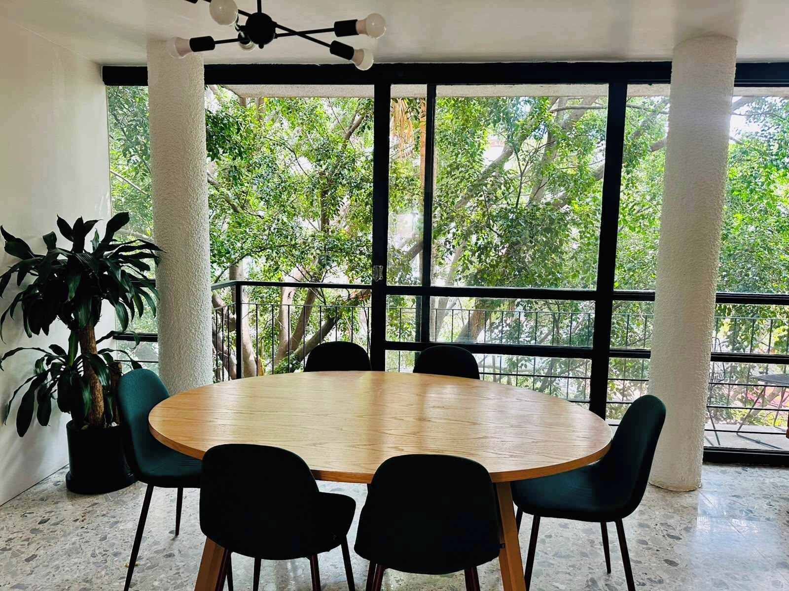A conference room with a round table and chairs, large windows with a view of trees, and a ceiling light fixture.
