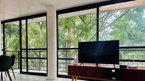 Un salón moderno con un televisor grande sobre un soporte de madera, una mesa de centro amarilla y vistas a árboles a través de amplios ventanales.