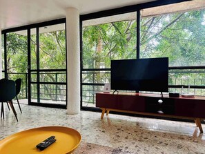 A modern living room with a large TV on a wooden stand, a yellow coffee table, and a view of trees through large windows. - Two-Level PH | Polanco · Near Masaryk · 4BR (Mexico City)