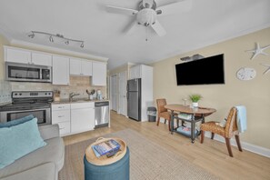 A modern kitchen with white cabinets, a microwave, stove, and a refrigerator. There's a dining area with a table and chairs, a TV mounted on the wall, and a ceiling fan. - Beachside Bliss - Beachfront Condo! (Hilton Head Island)