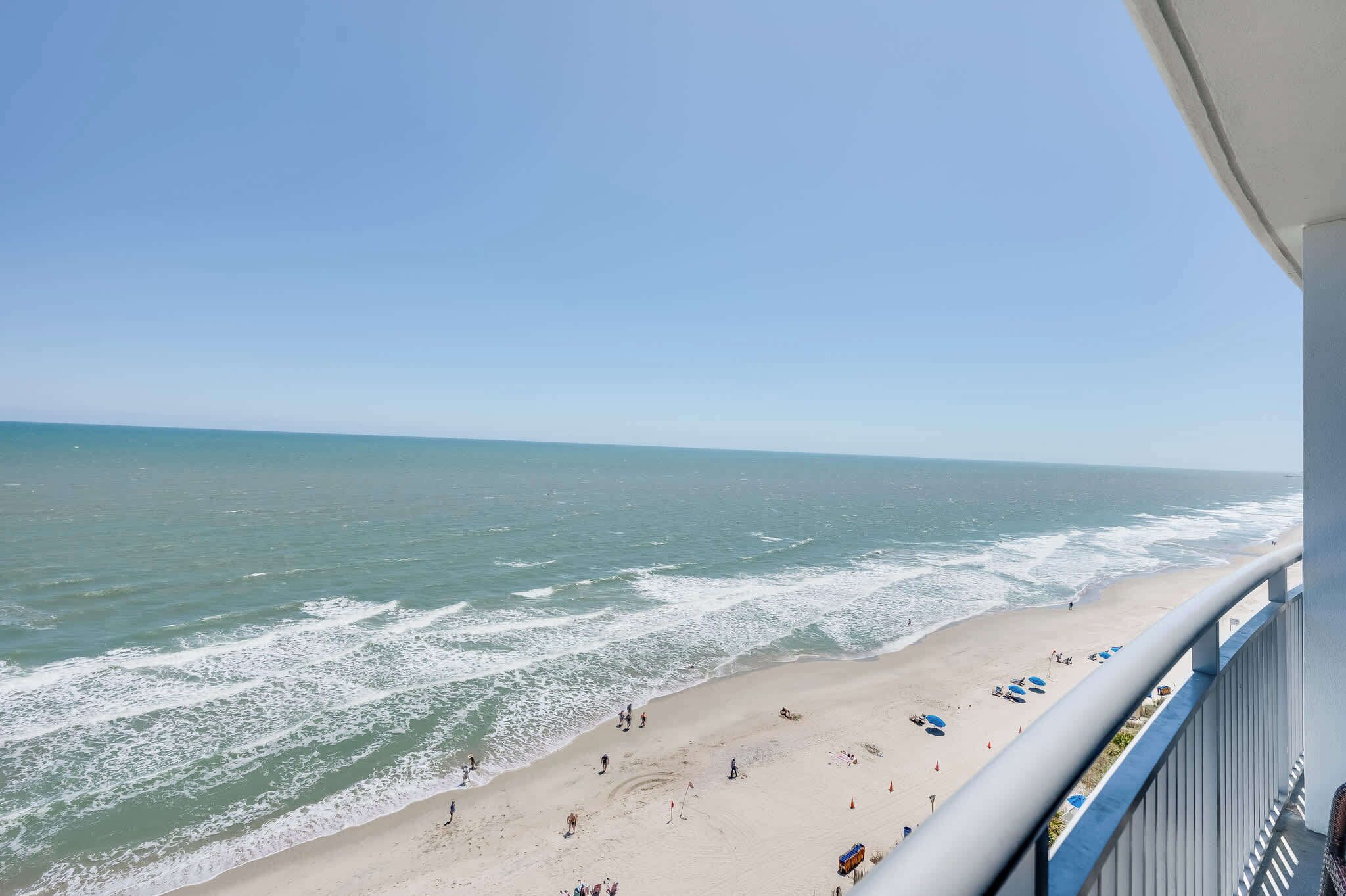 Une vue sur la plage depuis un balcon doté d’une balustrade, avec les vagues de l’océan et des personnes se promenant sur la plage.