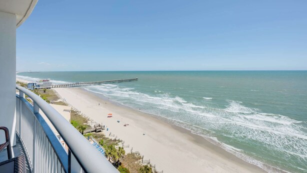 Ein Strand mit einem Steg, der sich ins Wasser erstreckt, ein klarer Himmel und ein Balkon mit Blick auf die Szene.