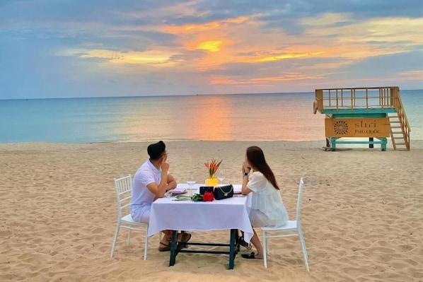 A couple dining at a beachside table with a sunset view, a lifeguard tower, and a bench in the background.