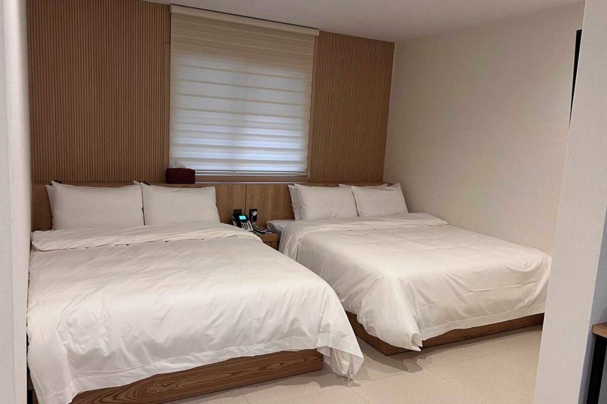 Quarto de hotel com duas camas, cabeceira de madeira, mesa de cabeceira com telefone e janela com persianas.