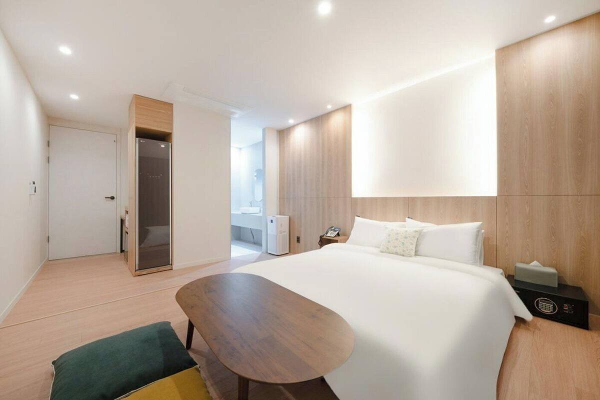Quarto de hotel moderno com uma cama grande, uma escrivaninha de madeira, um closet espaçoso e um banheiro visível através de uma porta aberta.
